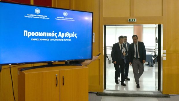 εικόνα που δείχνει σε πρώτο πλάνο μια μεγάλη οθόνη όπου αναγράφεται η φράση Προσωπικός Αριθμός από την ψηφιακή πλατφόρμα, ενώ στο φόντο διακρίνονται ο Μιχάλης Χρυσοχοΐδης αριστερά και ο Δημήτρης Παπαστεργίου δεξιά να προσέρχονται στη συνέντευξη Τύπου