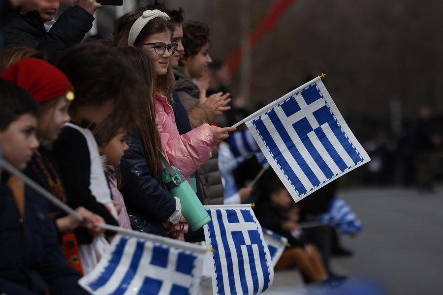 εικόνα που δείχνει παιδιά να παρακολουθούν τη στρατιωτική παρέλαση κρατώντας ελληνικές σημαίες