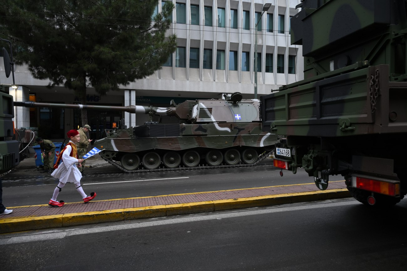 εικόνα που δείχνει μικρό μαθητή ντυμένο εύζωνα να περπατά ανάμεσα σε στρατιωτικά οχήματα λίγο πριν από την έναρξη της παρέλασης